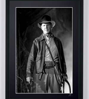 Black-and-white full-length studio photograph of Harrison Ford wearing Indiana Jones’s hat and leather jacket, with revolver and coiled whip in hand.