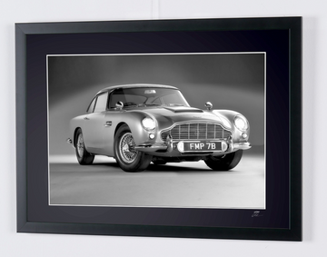 James Bond 007 : Goldfinger (1964) — Aston Martin DB5, coche de tres cuartos delantero sobre fondo liso, con matrícula FMP 7B visible, fotografía de cine enmarcada en blanco y negro, marco visto en ángulo desde la izquierda.