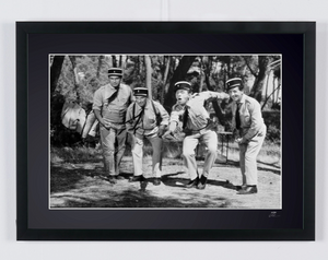 Black-and-white full-length photograph of four summer-uniformed gendarmes playing pétanque on a sunlit dirt court among pine trees.
