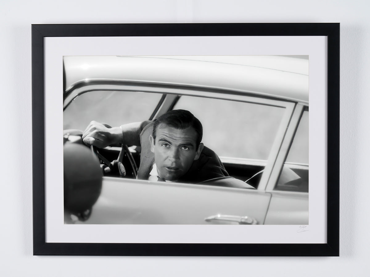 James Bond 007: Goldfinger (1964) - Sean Connery achter het stuur van de Aston Martin DB5, tussen twee opnames — ingelijste filmfoto
