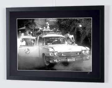 Ghostbusters (1984) — Ecto-1, Ecto-1 car seen from the front, with a door open and smoke near the ground, framed cinema photograph in black and white, frame viewed at an angle from the left.
