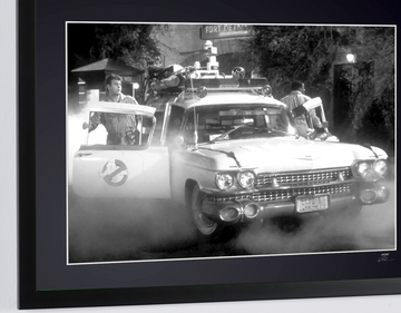 Ghostbusters (1984) — Ecto-1, Ecto-1 car seen from the front, with a door open and smoke near the ground, framed cinema photograph in black and white, close-up view of the photograph and frame.