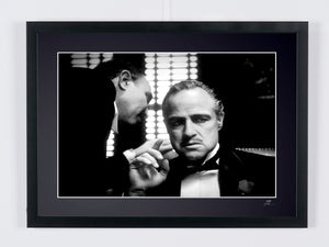 Black-and-white photograph of Marlon Brando in a tuxedo, bow tie and boutonnière as a man leans in to speak at his ear.